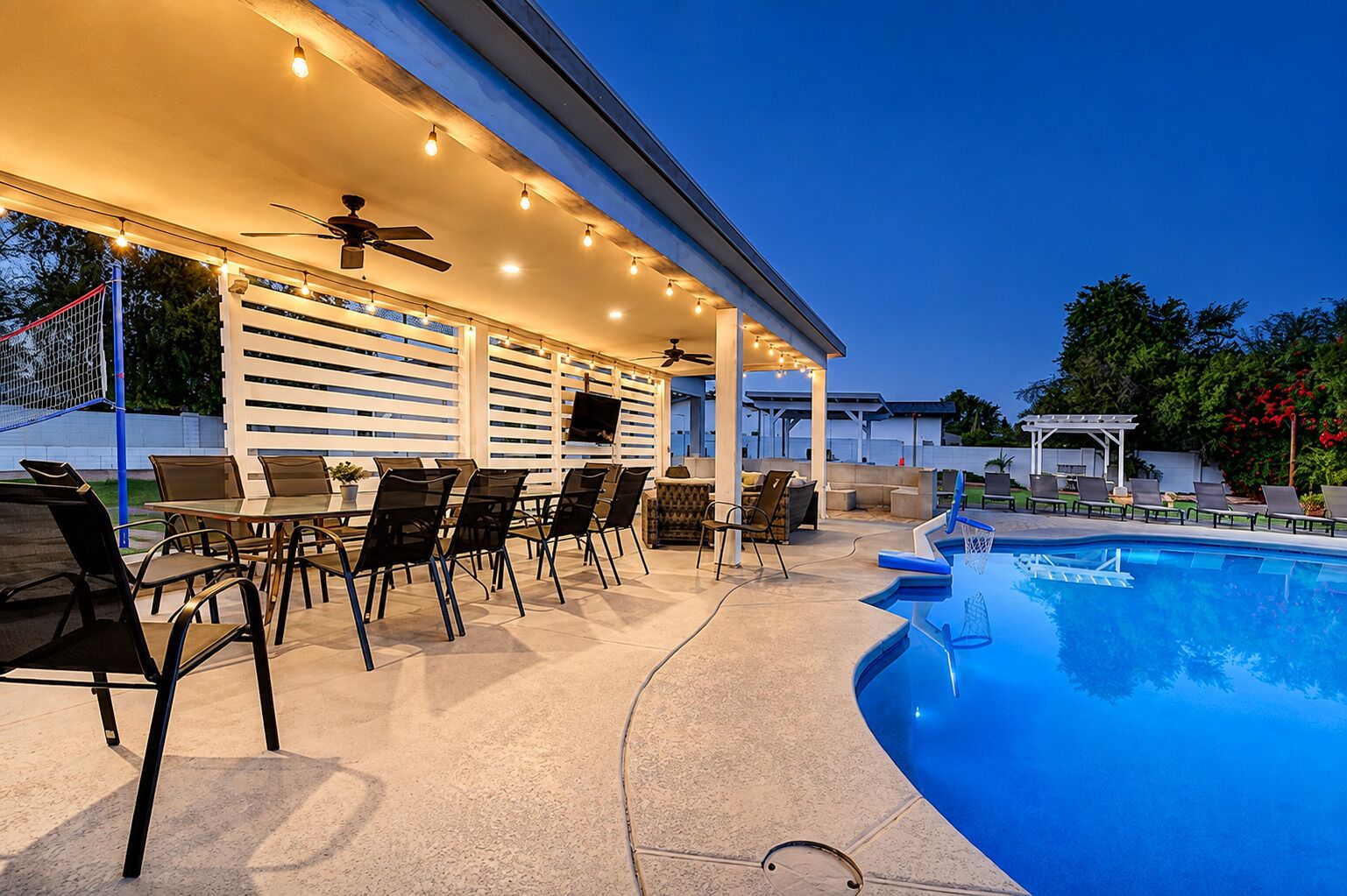 Un área de piscina exterior con zona de estar techada, sillas y un ventilador de techo.