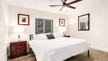 Un dormitorio con una cama, dos mesitas de noche, un ventilador de techo, una ventana con persianas y cuadros en la pared.