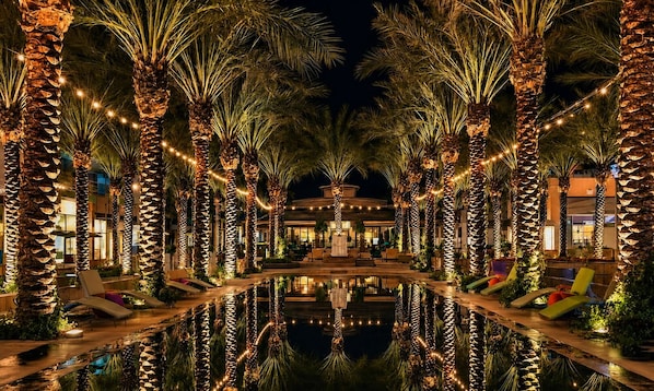 A poolside area with palm trees and lounge chairs, reflecting a building in the background.