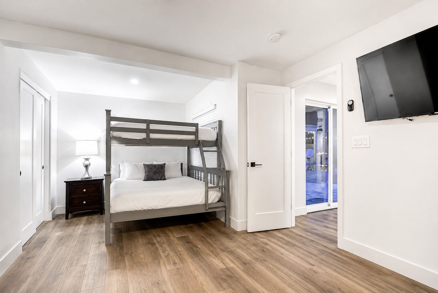 Un dormitorio con una cama litera, una mesita de noche con lámpara, un televisor montado en la pared y una puerta que da a otra habitación.