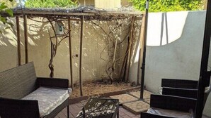 Area patio esterna con una pergola, due poltrone in vimini, un tavolino e un ombrellone grande che offre ombra.