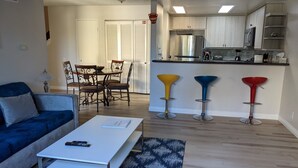 Un soggiorno con un divano blu, un tavolino da caffè bianco e un tappeto decorato. C'è una zona pranzo con tavolo e sedie, e una cucina con frigorifero, piano cottura e sgabelli da bar.