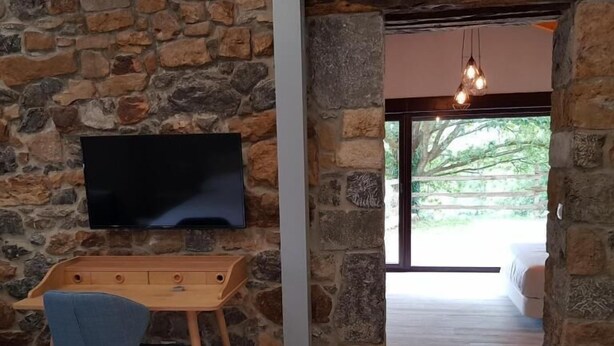 Habitación con paredes de piedra, un escritorio de madera, una silla, un televisor de pantalla plana y un acceso a un balcón con vistas.