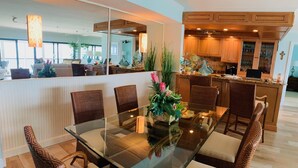 Un espace repas moderne avec une table en verre entourée de chaises en osier, une cuisine en arrière-plan et un grand miroir qui reflète l’ensemble de la pièce.
