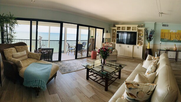 Un vaste salon doté d’une grande porte-fenêtre coulissante donnant sur un balcon avec vue sur la mer. On y trouve un canapé confortable, une table basse en verre et un téléviseur fixé à un meuble.