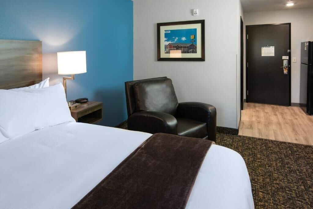A hotel room with a bed, a chair, a desk with a lamp, a framed picture on the wall, and a door.