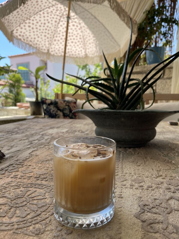 A glass of iced coffee on a textured table with a potted plant and a patterned umbrella in the background. - Afet Inn Hotel (Cesme)