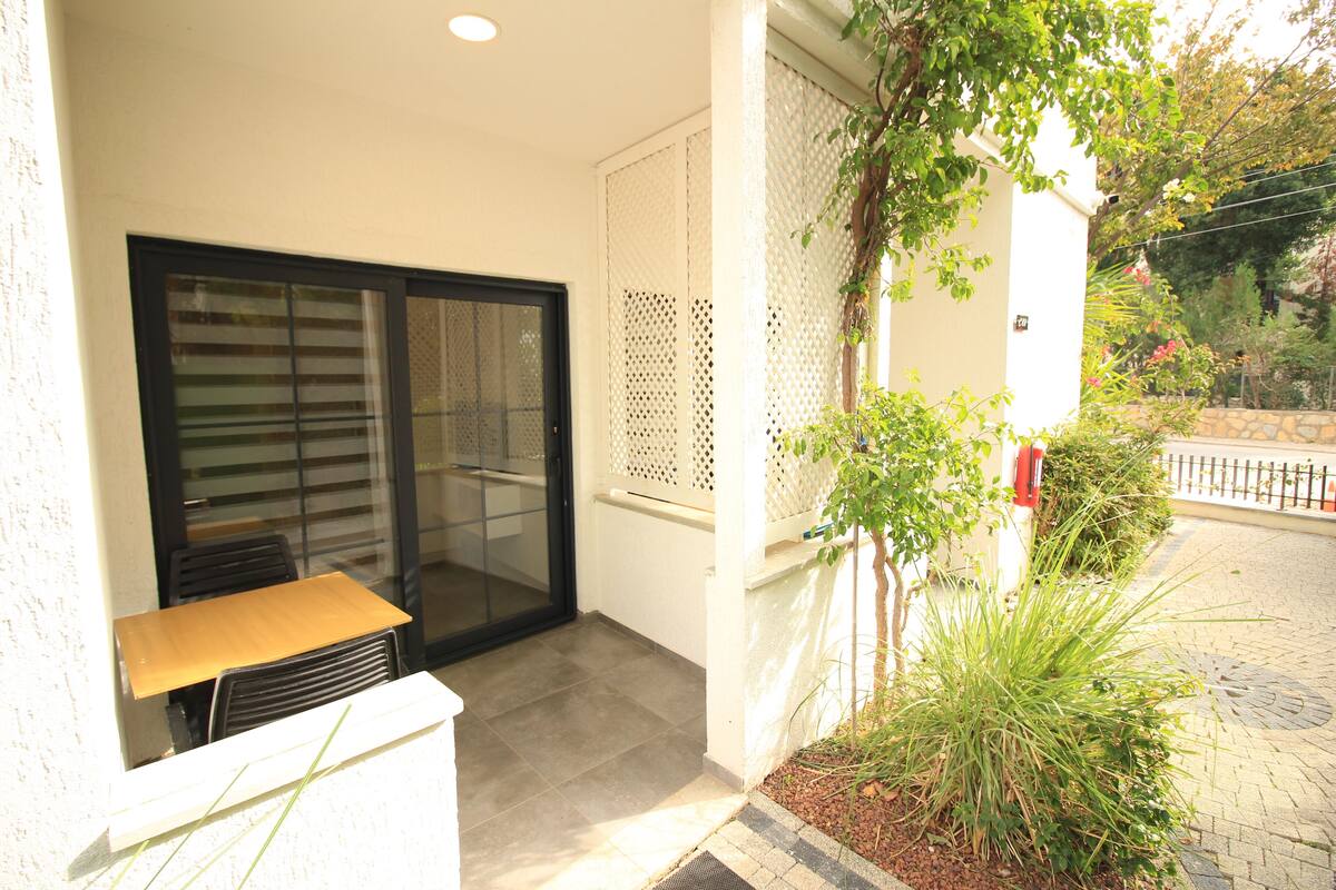 A small balcony with a wooden table and chairs, a sliding glass door, and a potted plant.