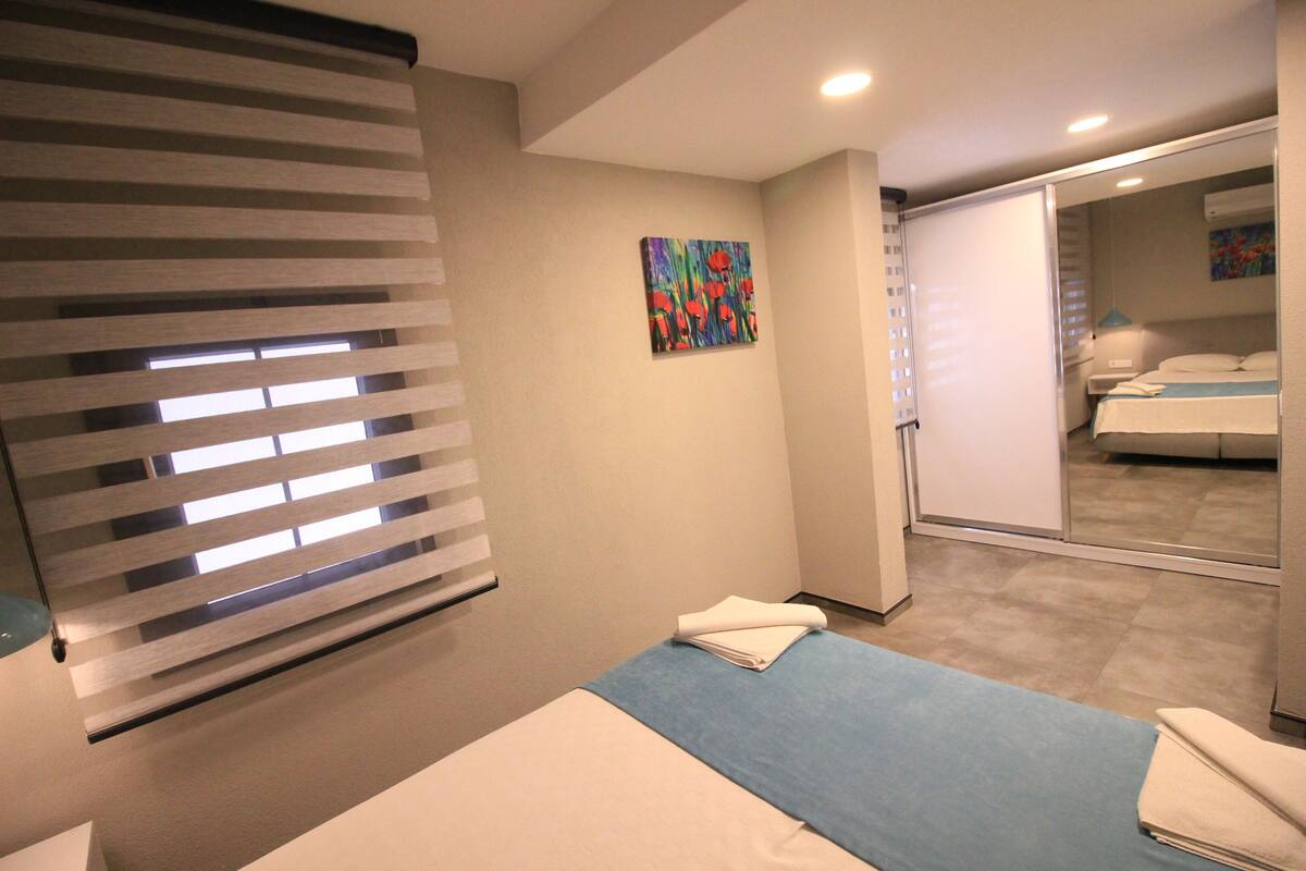 A hotel room with a bed, a window with blinds, a painting on the wall, and a bathroom visible through a glass door.