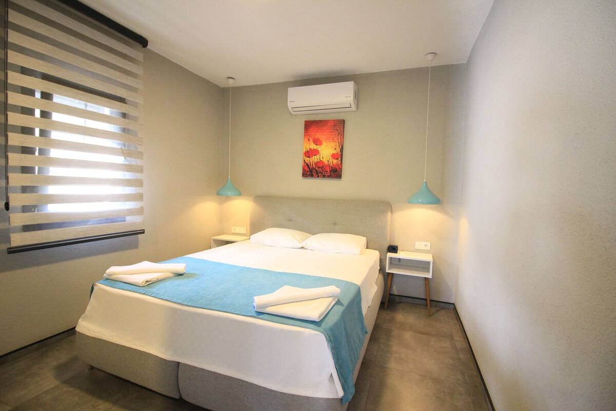 A hotel room with a bed, two bedside tables, an air conditioner, a painting, and a window with blinds.