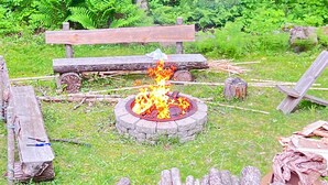 Ein Lagerfeuer, umgeben von Holzscheiten und einer hölzernen Bank, inmitten eines Waldes.