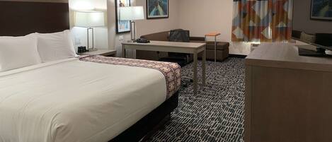A hotel room with a large bed, a desk, a chair, a TV, and a patterned carpet.