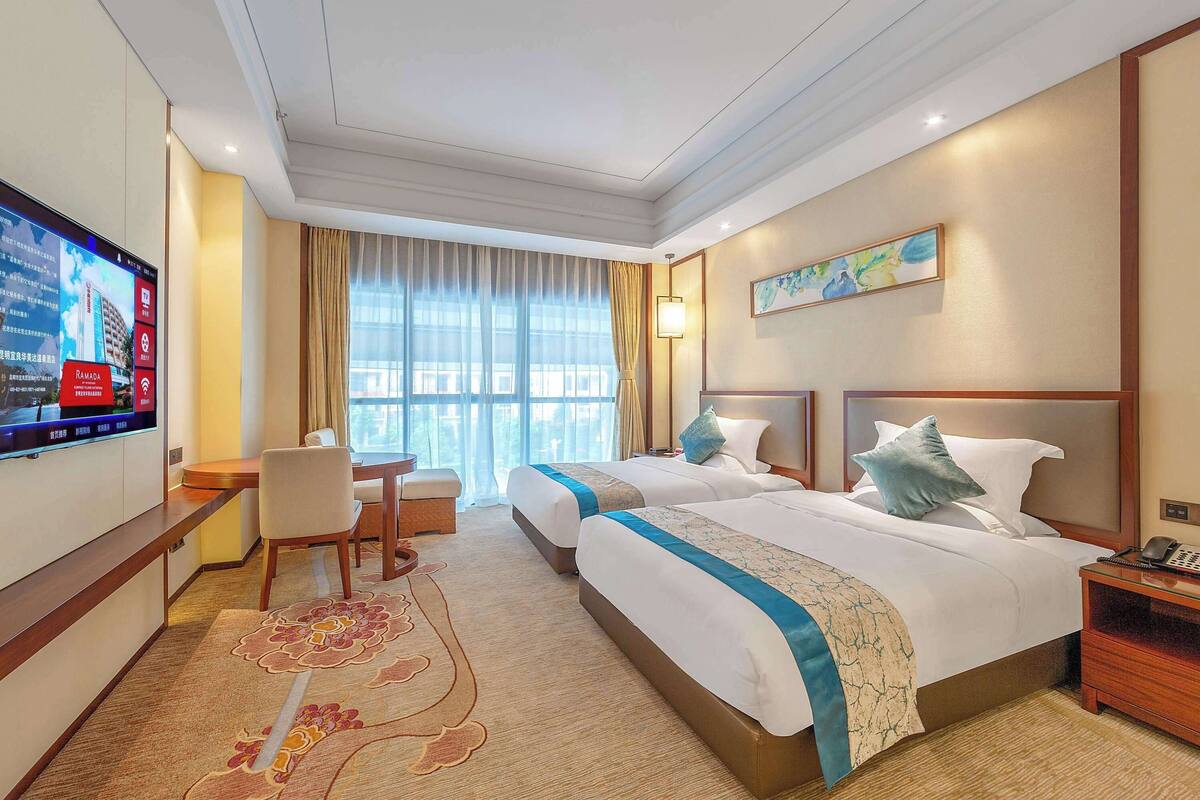 A hotel room with two beds, a desk, a TV, and a large window with curtains.