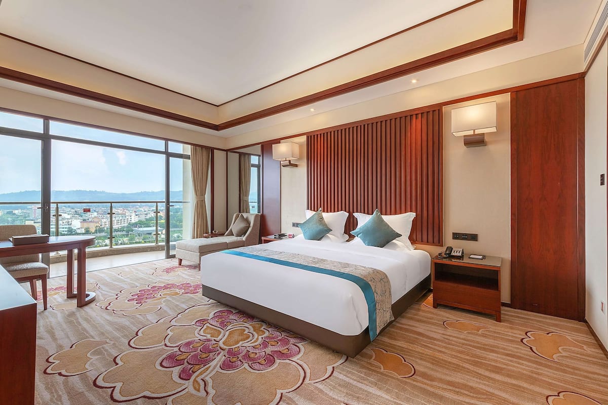 A hotel room with a large bed, a desk, a chair, a nightstand, and a view of the city.