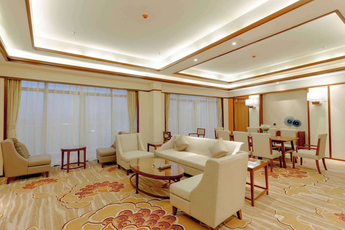 A spacious hotel room with a large sofa, armchairs, a coffee table, and a dining area with chairs and a table.