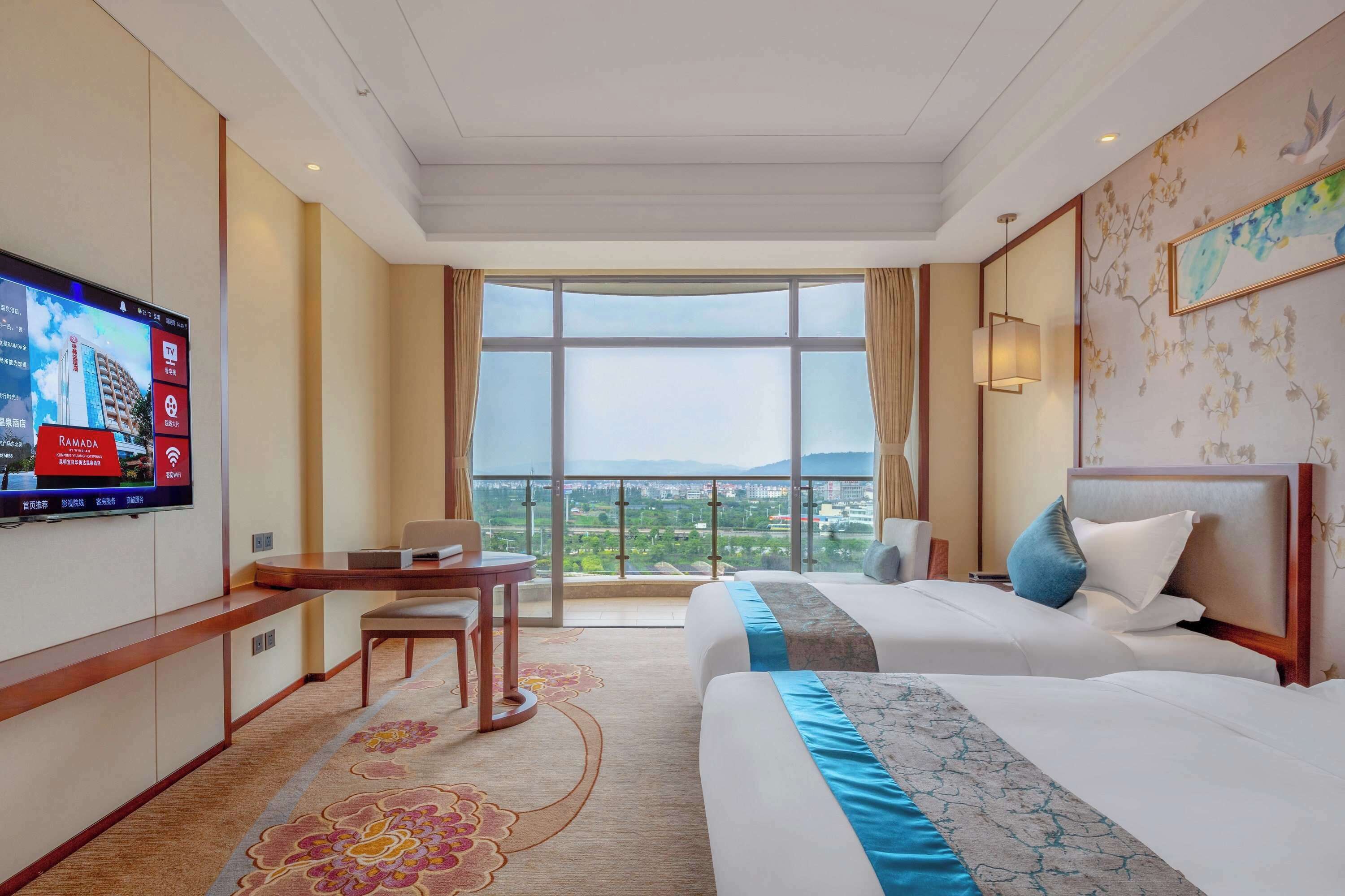 A hotel room with two beds, a desk, a TV, and a large window with a view.