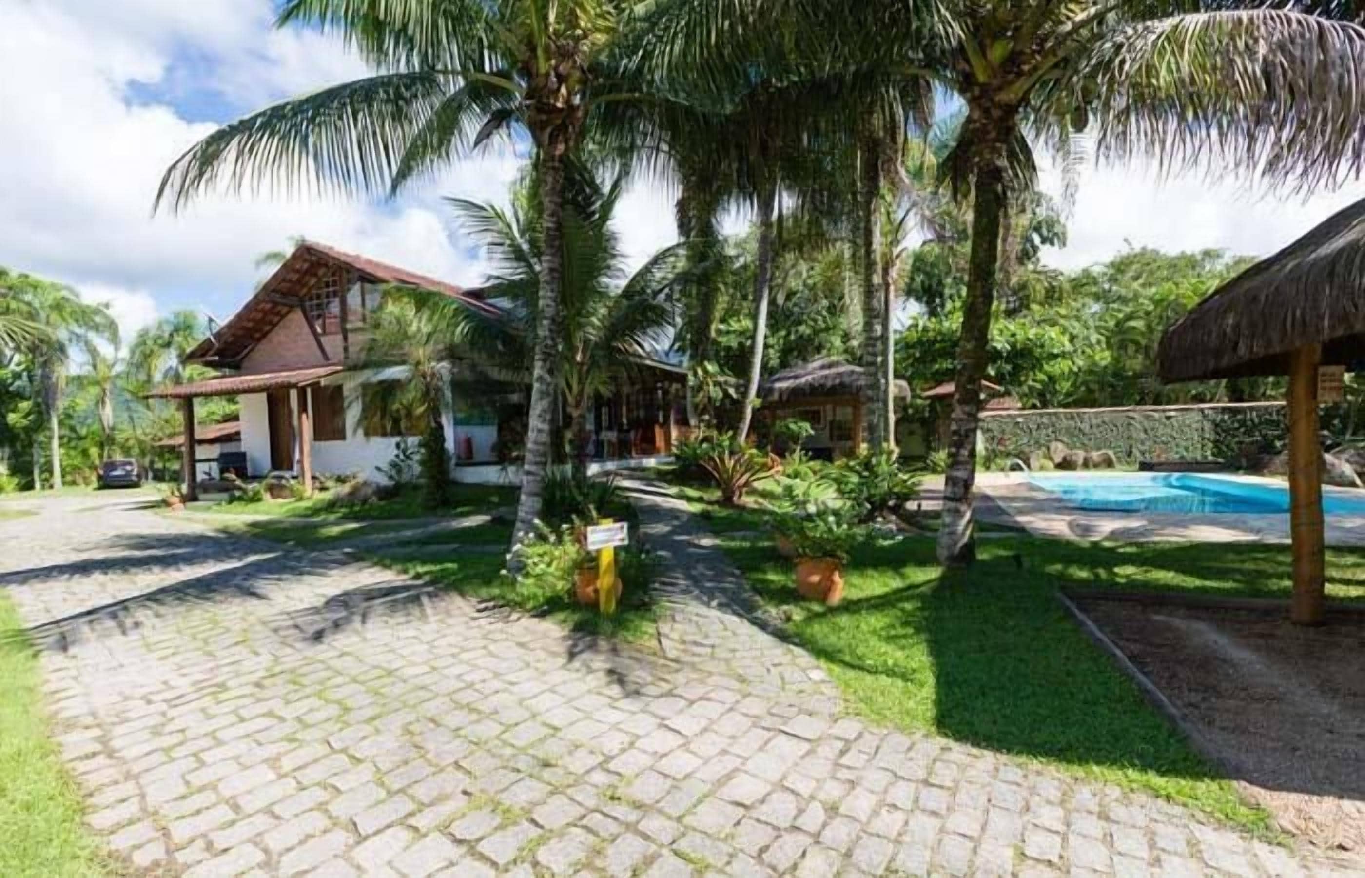 A tropical resort with a paved walkway, palm trees, a pool, and a thatched-roof structure.