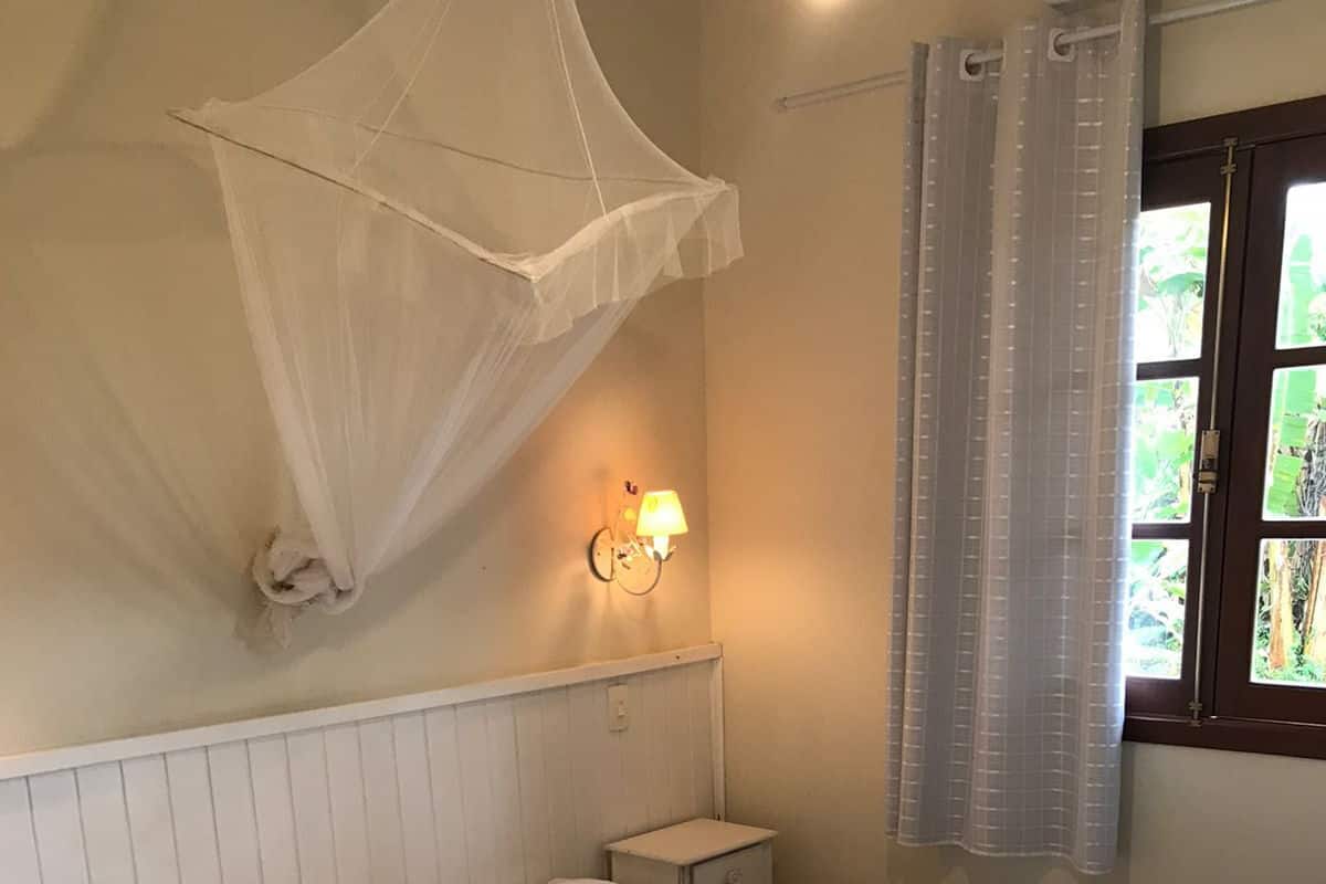 A bed with a canopy, a bedside table, a nightstand, a wall-mounted lamp, and a window with curtains.