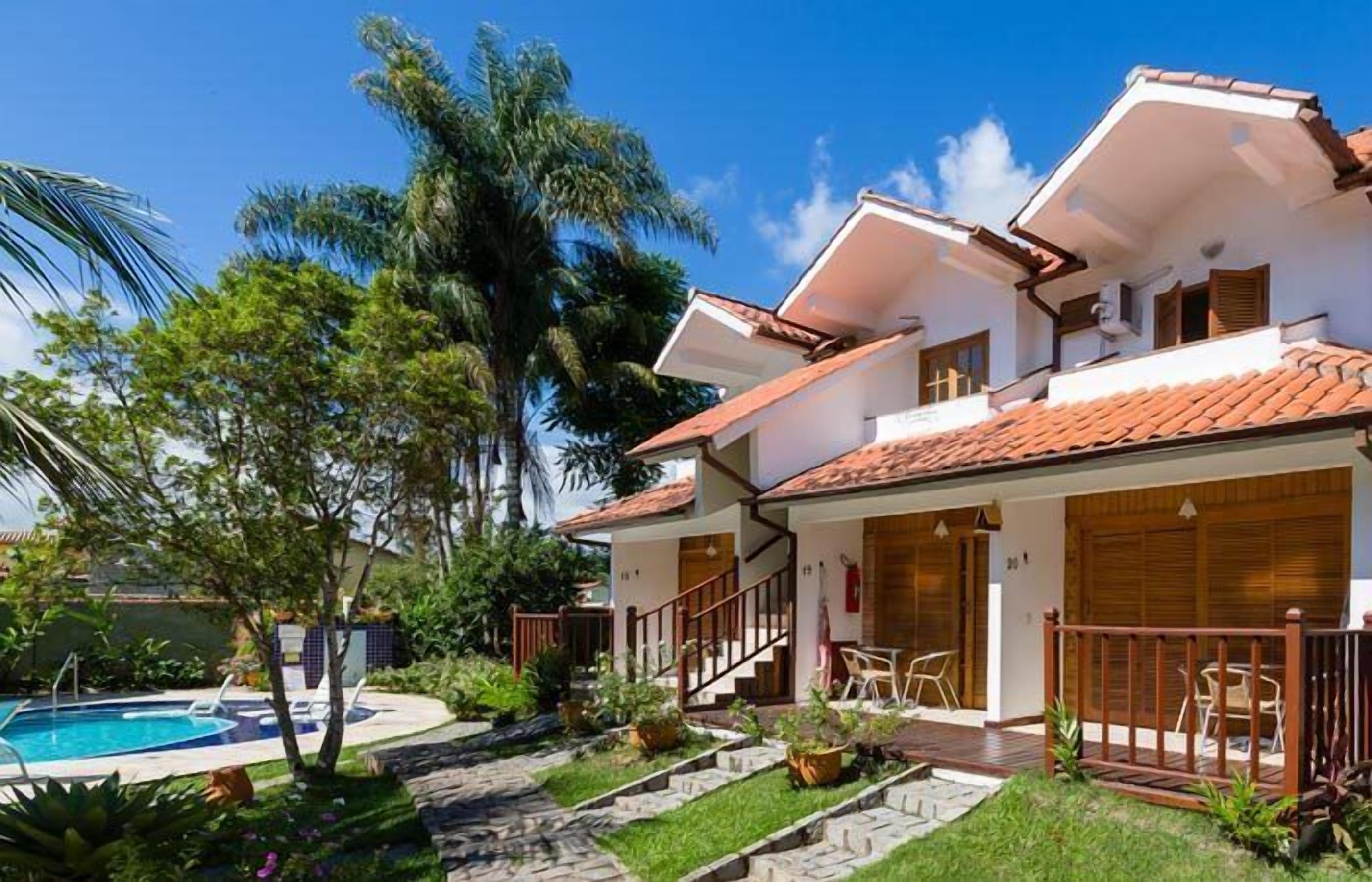 A two-story house with a swimming pool, surrounded by lush greenery and palm trees.