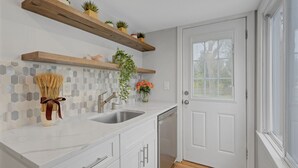 Uma cozinha com armários brancos, bancada de mármore, lava-louças embutida, prateleiras de madeira com plantas e painel de azulejos hexagonais.