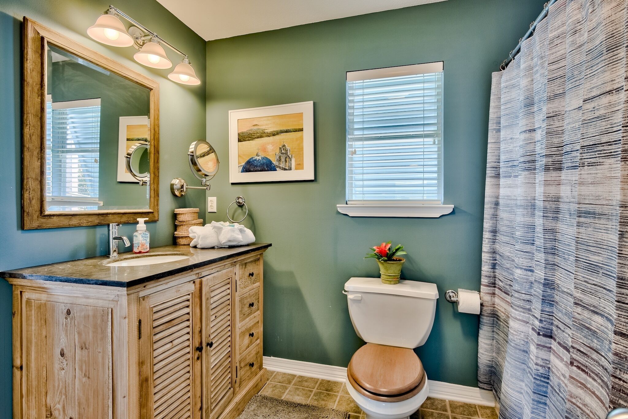 A bathroom with a wooden vanity, a large mirror, a toilet with a wooden seat, a framed picture on the wall, and a shower curtain.