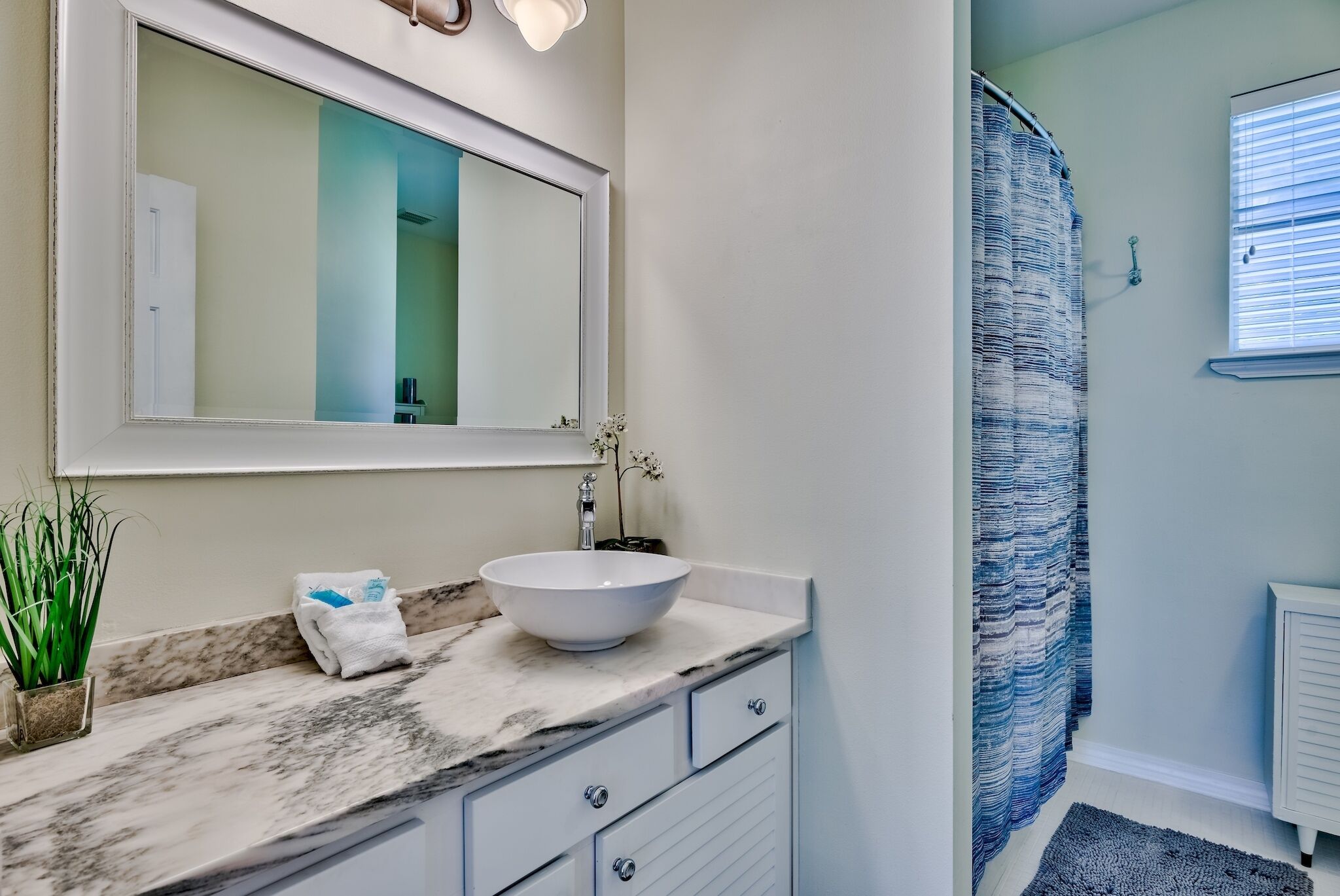 Un baño con encimera de mármol, un lavabo blanco, un espejo, una planta en maceta y una cortina de ducha.