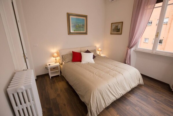 A bedroom with a bed, two bedside tables with lamps, a radiator, and a window with curtains.