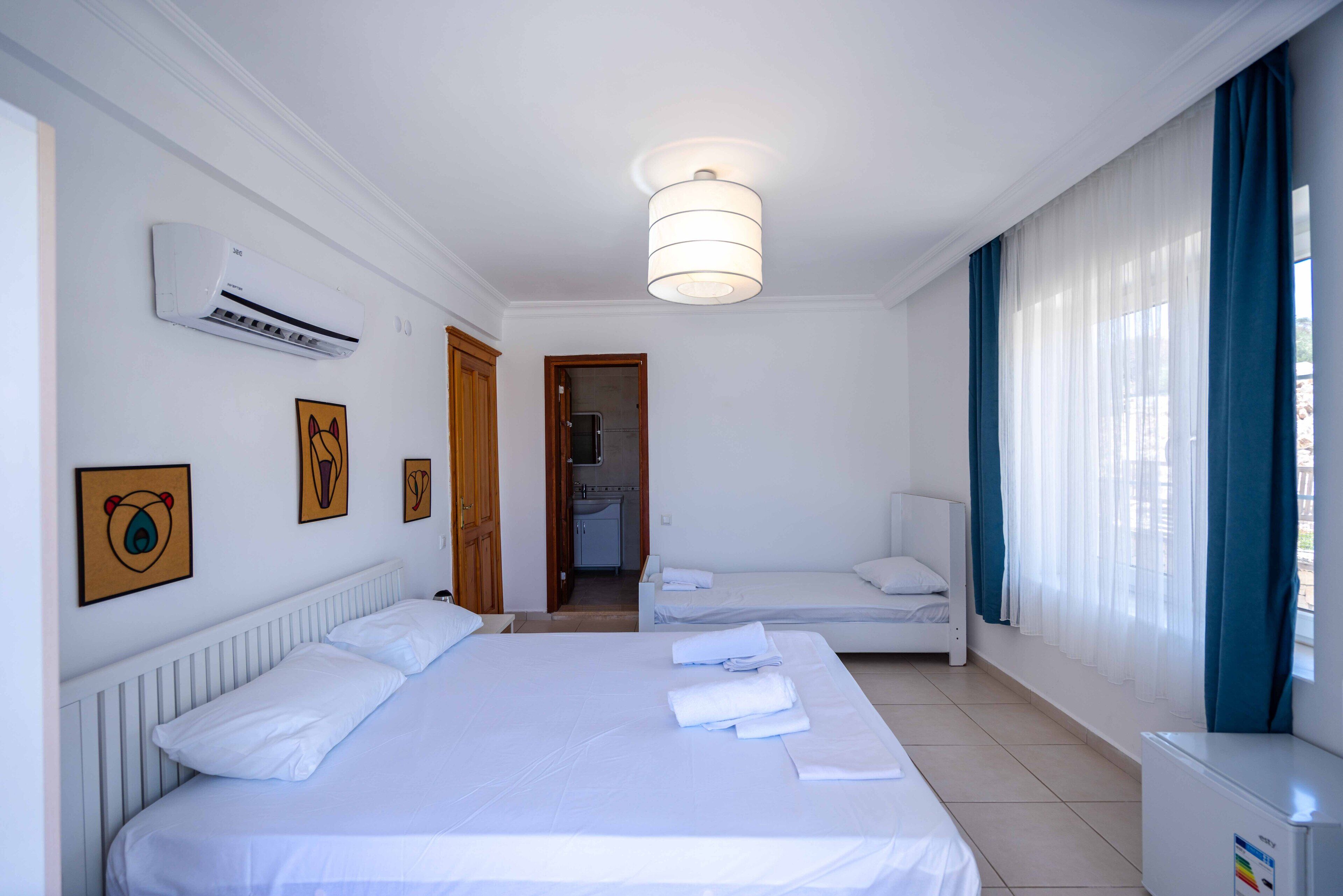 Habitación de hotel con una cama grande, una cama más pequeña, un equipo de aire acondicionado, una lámpara de techo y vistas al exterior a través de una ventana con cortinas.