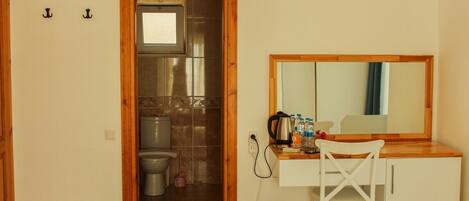 A compact room with a wooden door leading to a bathroom, a small desk with a chair, a mirror, and a trash bin.