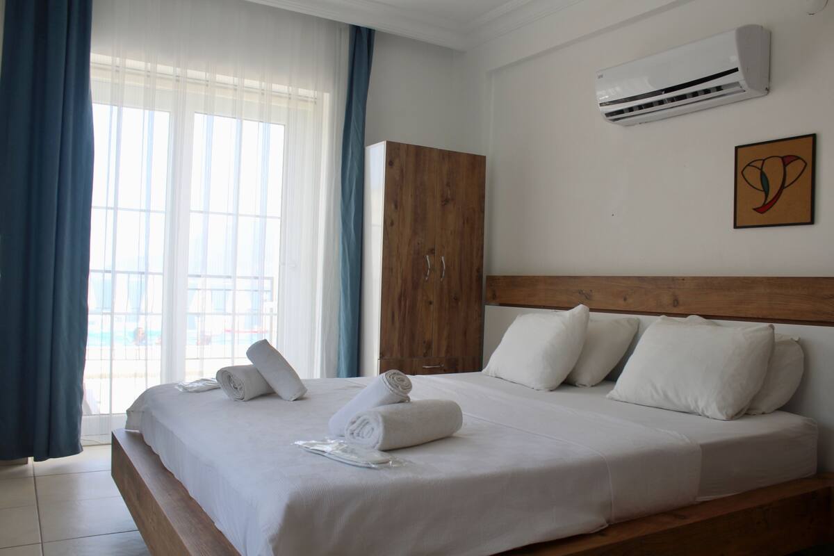 A hotel room with a large bed, a wooden wardrobe, an air conditioning unit, and a window with curtains.