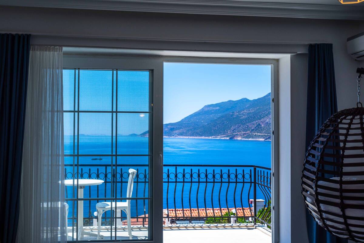 A room with a large sliding glass door leading to a balcony with a view of the sea and mountains.