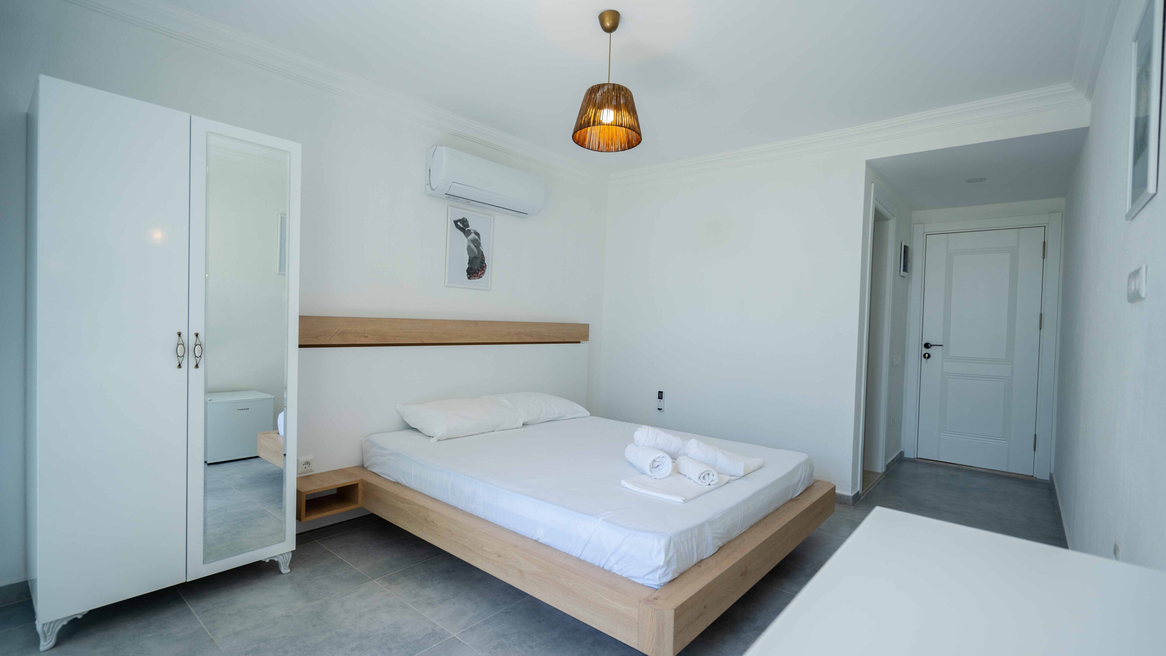 A modern bedroom with a wooden bed frame, a white mattress, and a bedside table. There is a wardrobe with a mirror, an air conditioning unit on the wall, and a hanging light fixture.