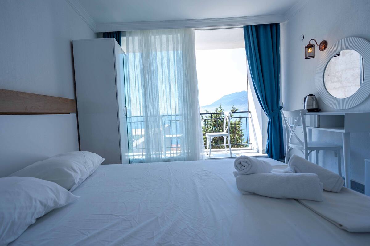 A hotel room with a large bed, a bedside table with a mirror, a view of the balcony, and a mountain in the background.