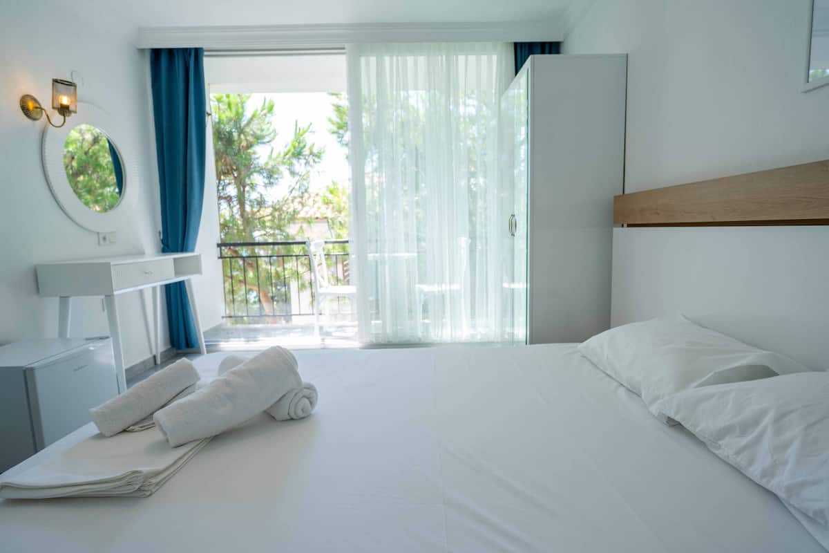 A hotel room with a bed, a desk, a chair, and a balcony with a view of trees.