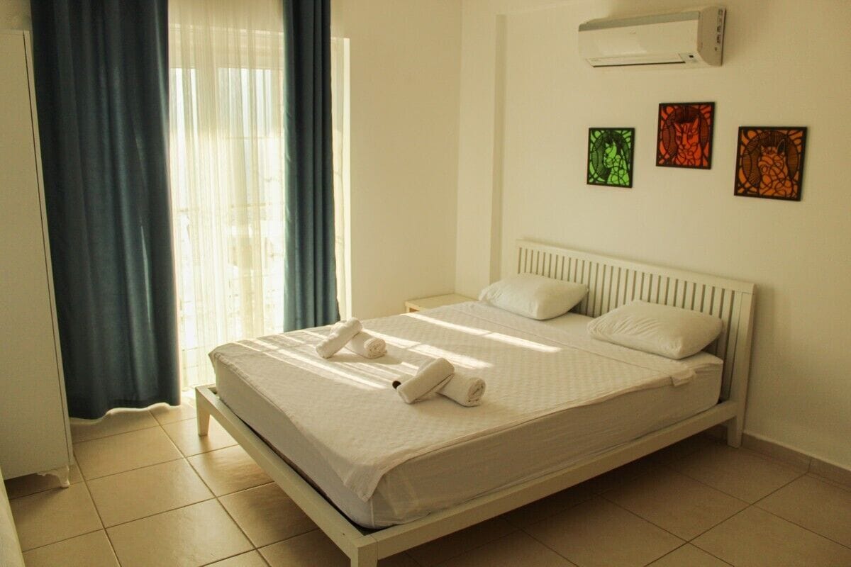 Una cama bien hecha con sábanas blancas, cabecera blanca y aire acondicionado instalado en la pared.