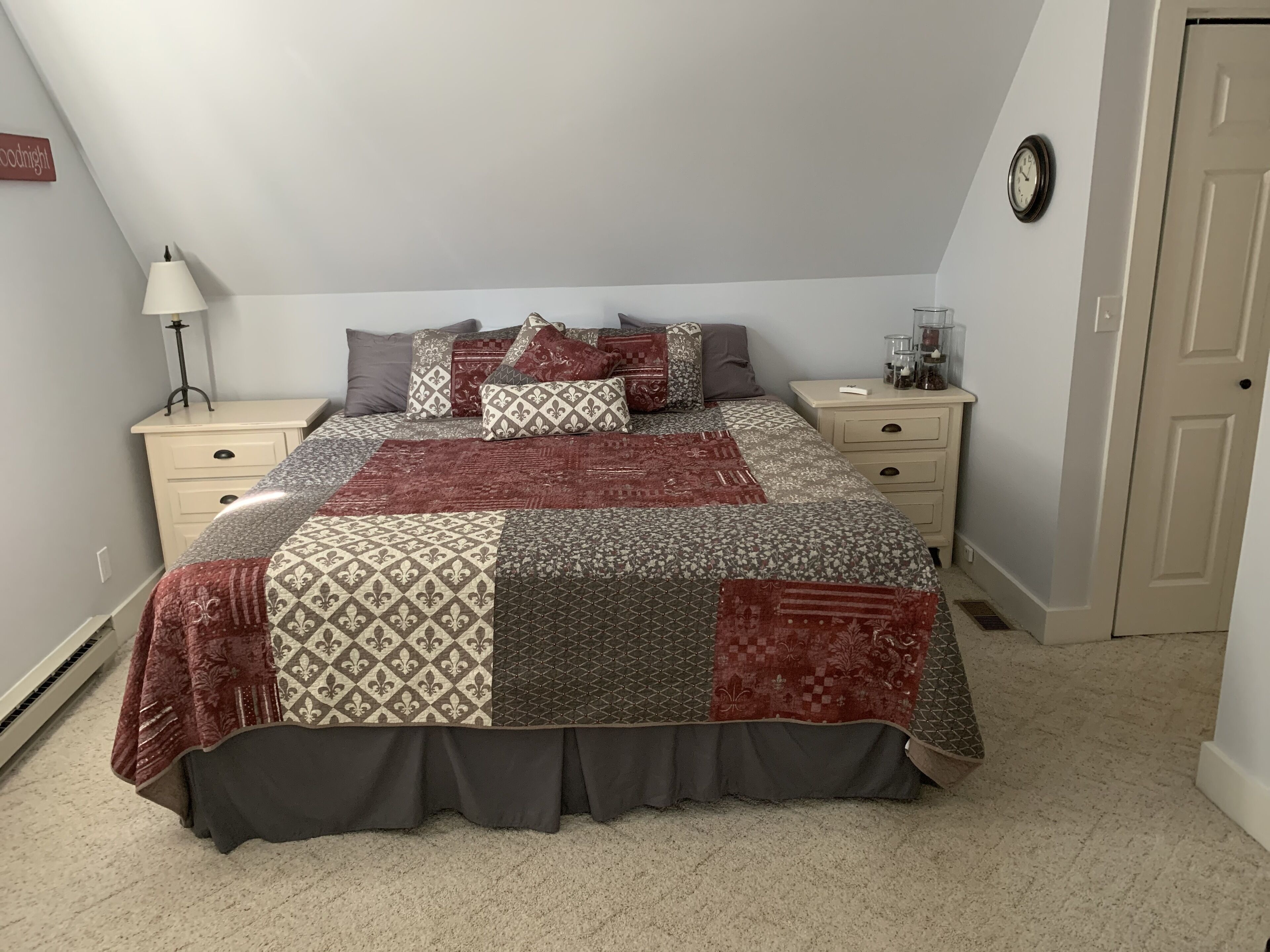 Une chambre à coucher avec un grand lit, deux tables de chevet, une lampe, une horloge et une porte.