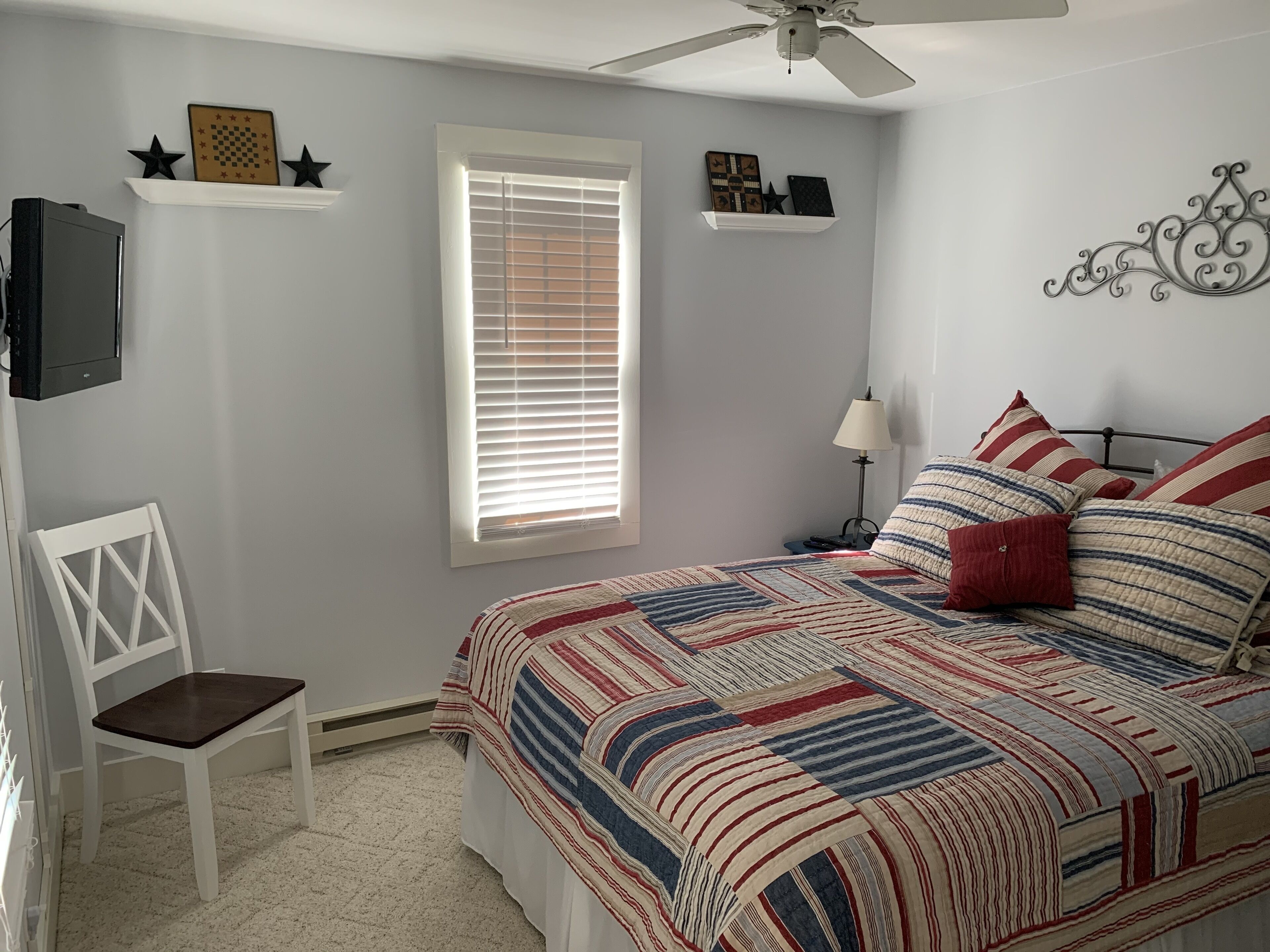 Un dormitorio con una cama, una silla, un ventilador de techo, una ventana con persianas y decoración en las paredes.