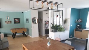 A modern living room with a wooden dining table, colorful chairs, a foosball table, and a kitchen area with a blue accent wall.