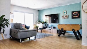 A modern living room with a grey sofa, a coffee table, and a foosball table. There is a TV mounted on the wall and a window providing natural light.