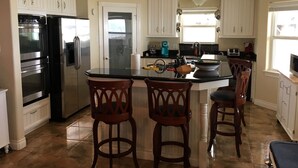 Une cuisine avec des meubles blancs, un réfrigérateur noir, un îlot central avec une plaque de cuisson en verre et des tabourets de bar, et une fenêtre qui laisse entrer la lumière naturelle.