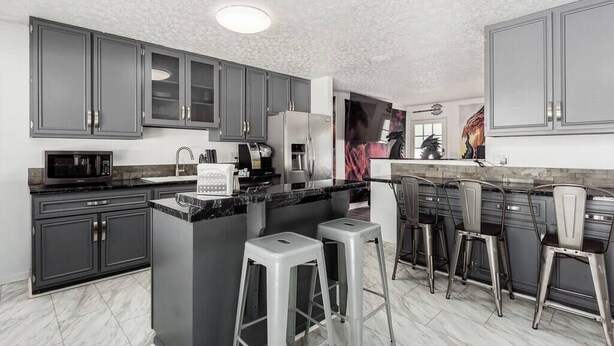 A modern kitchen with grey cabinets, a central island with stools, a microwave, and a coffee machine. - ★Dragon’s Lair★Private Home w/ Arcade & Minigolf★ (Westerville)