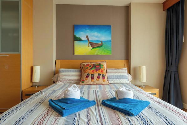 A hotel room with a bed, a painting of a boat on water, a patterned pillow, and two towels on the bed.