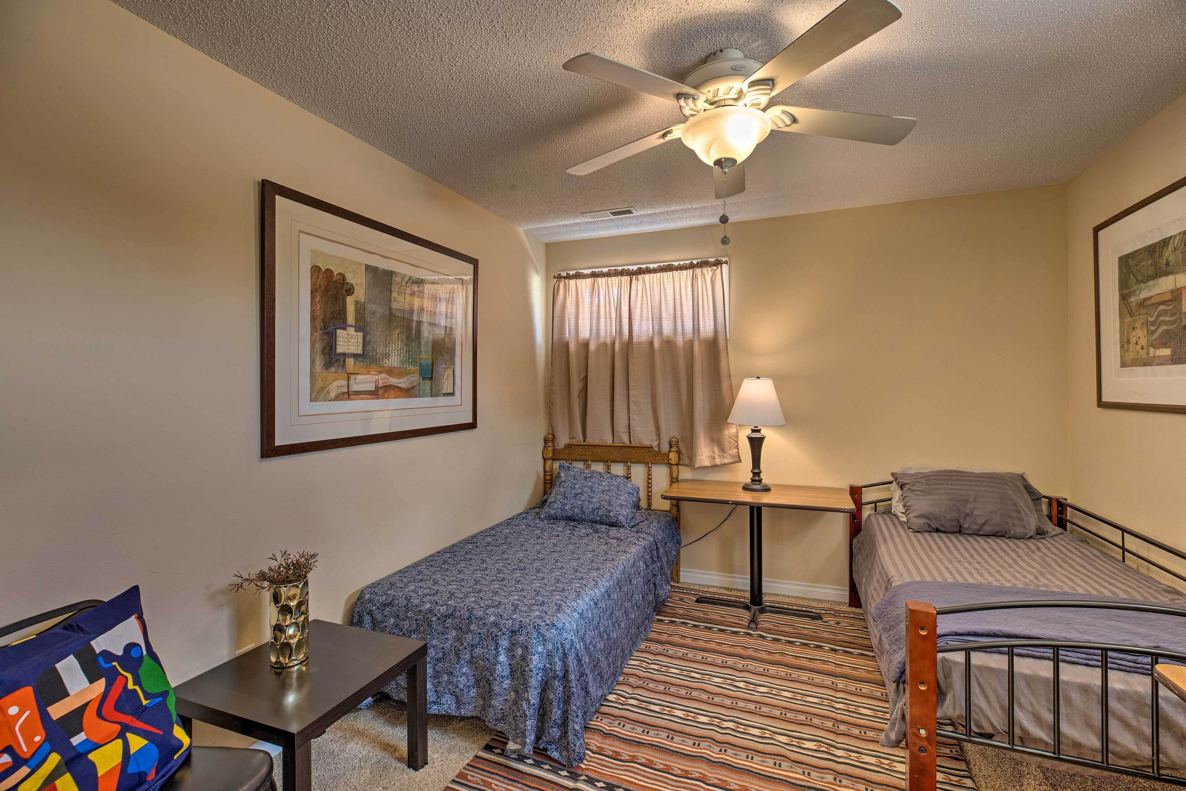 A bedroom with two single beds, a ceiling fan, a bedside table with a lamp, a small table with a vase, and a colorful bag on a chair.