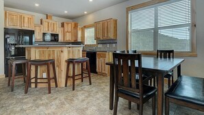 Une cuisine avec des meubles de rangement en bois, un îlot central avec des tabourets, une table à manger avec des chaises et une grande fenêtre avec des stores.