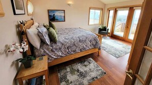 Un dormitorio con una cama grande, una mesita de noche con lámpara, una silla pequeña y vistas al exterior a través de un ventanal.