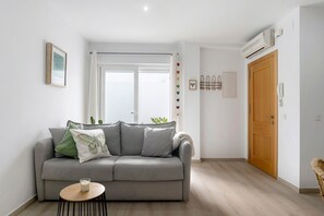 En stue med en grå sofa, et lille rundt bord, et vindue med gardiner, en trædør og en aircondition.
