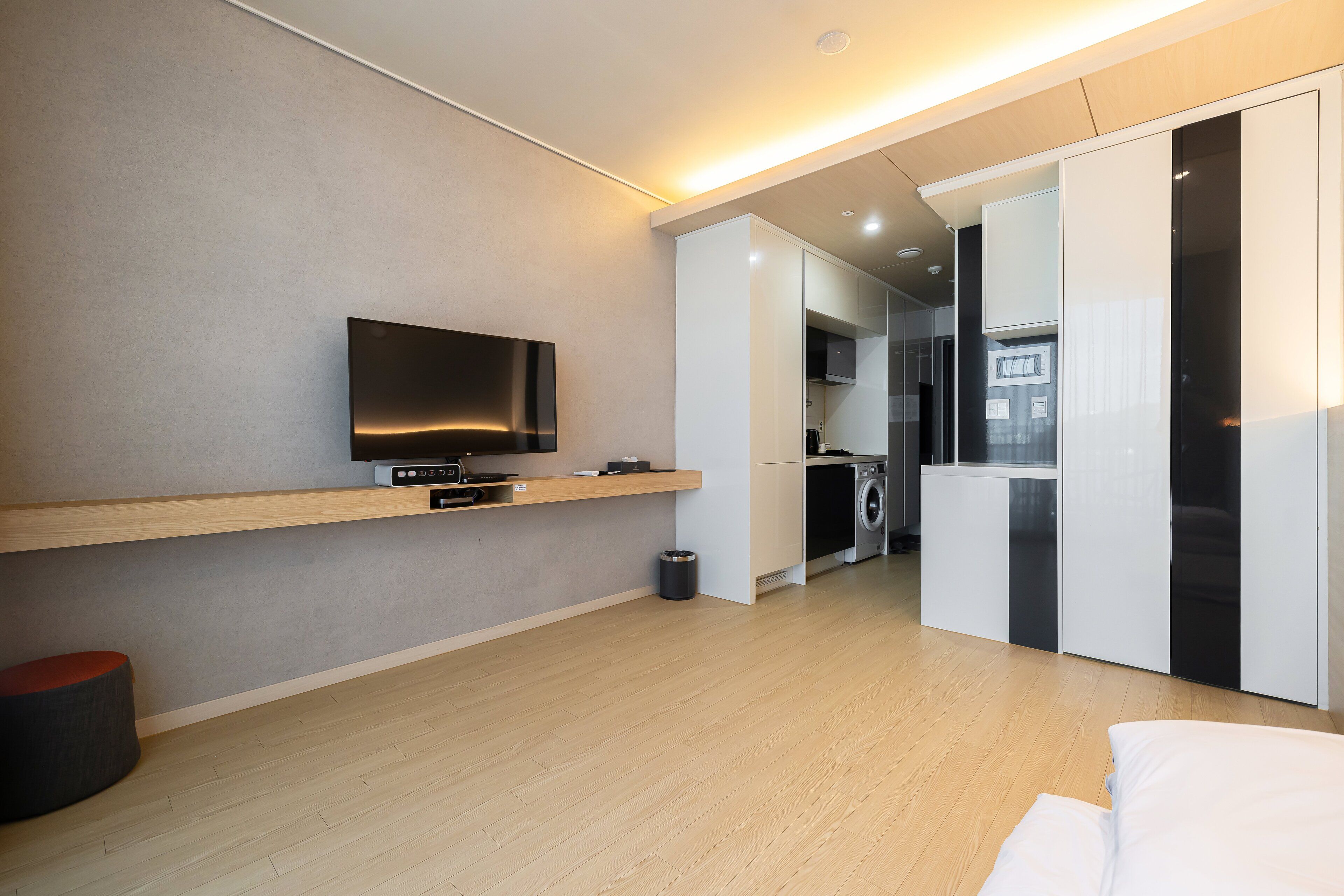 A modern, well-lit room with a flat-screen TV mounted on a wooden shelf, a compact kitchen area with a washing machine, and a large wardrobe.