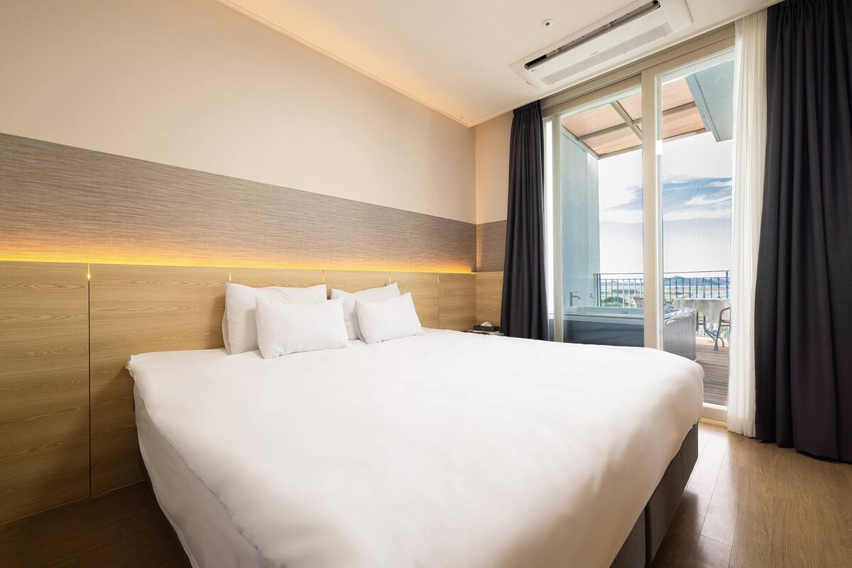 A large bed with white linens, a wooden headboard, and a view of a balcony with outdoor furniture.