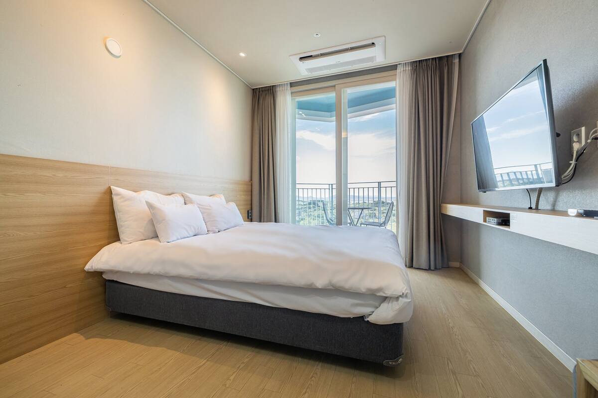 A modern hotel room with a large bed, a flat-screen TV mounted on the wall, and a balcony with a view.