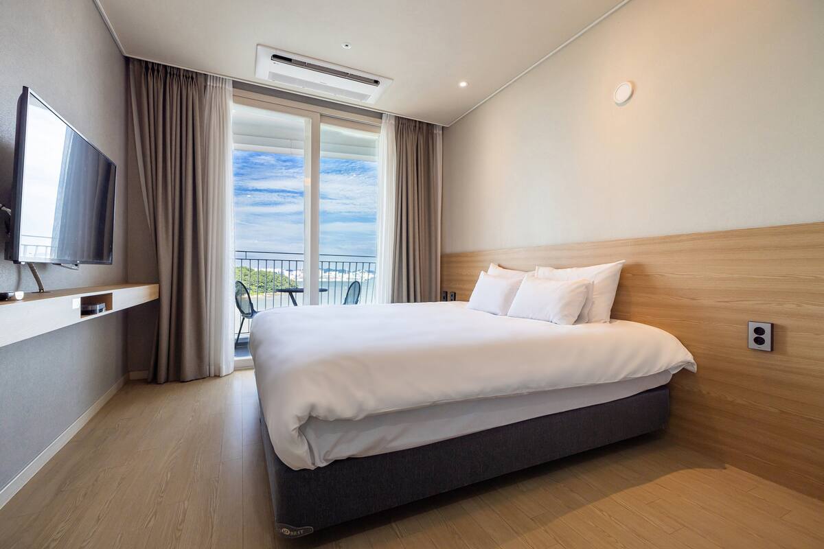 A modern hotel room with a large bed, a flat-screen TV, a balcony with a view, and a wooden headboard.